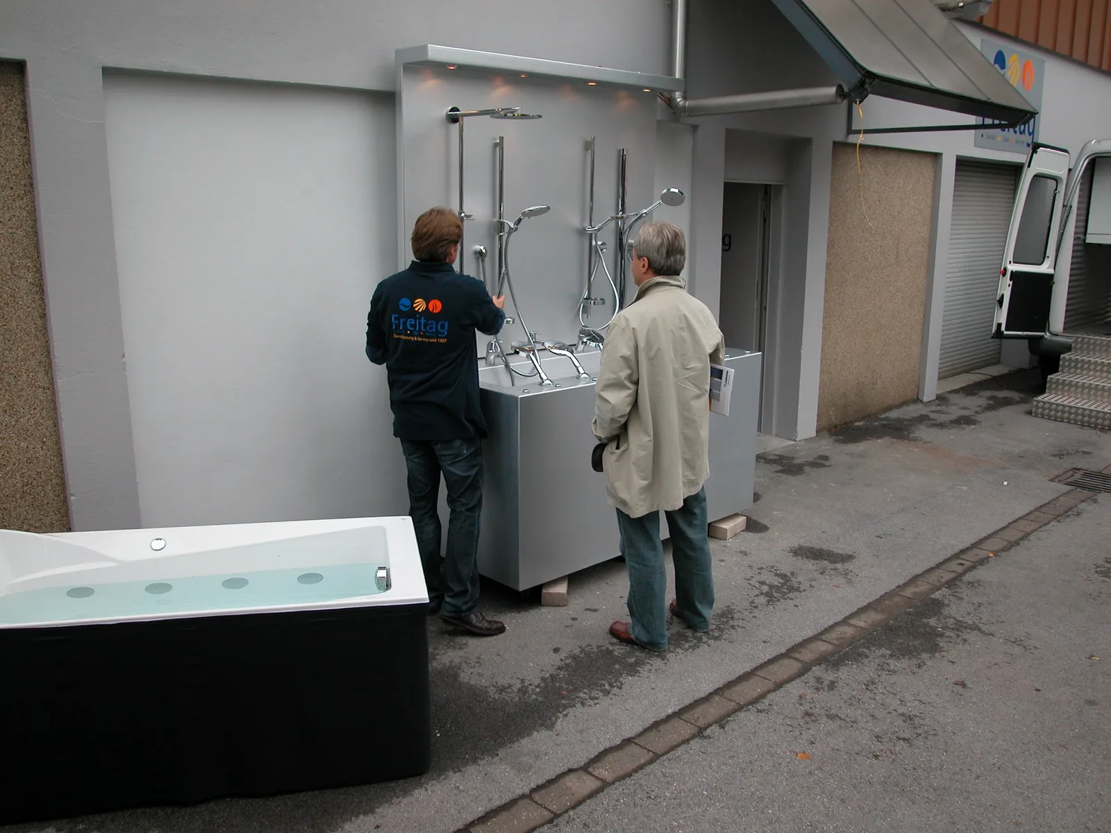 Mitarbeiter in Freitag-Jacke zeigt einem Besucher Duschpaneele im Hof, davor eine Whirlwanne